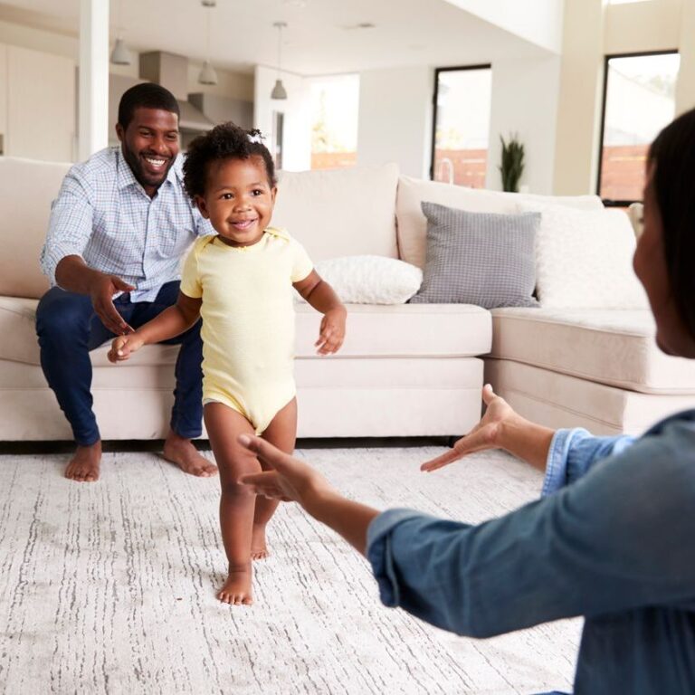 A parent and toddler experiencing a first moment together in a cozy home setting. The toddler is taking their first steps, with the parent close by, cheering and encouraging. The room is warm and welcoming, filled with toys and family photos, creating a heartwarming and joyful atmosphere.