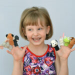 A young girl smiling and playing with colorful finger puppets in a bright room with toys and educational materials.