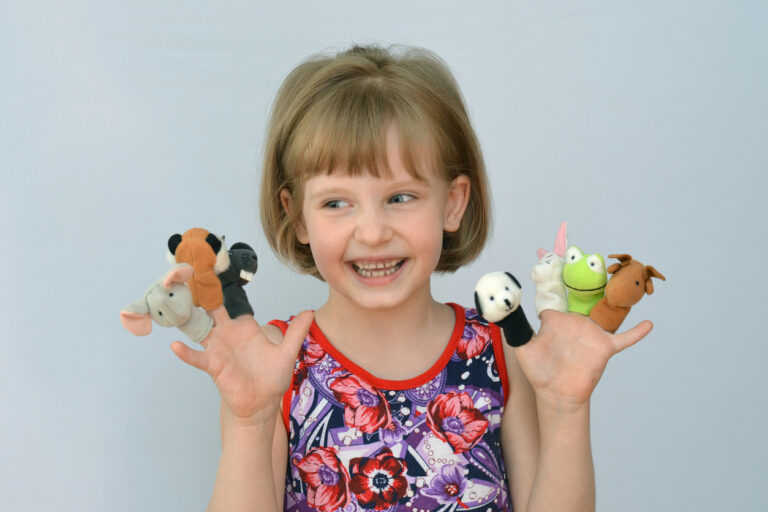 A young girl smiling and playing with colorful finger puppets in a bright room with toys and educational materials.